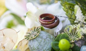 Zwei Eheringe ruhen auf einer strukturierten Oberfläche, umgeben von grünem Laub und weißen Blumen, und schaffen so eine natürliche und romantische Kulisse. Ideal für alle, die gerade mitten in der Hochzeitsplanung stecken, denn der Hintergrund ist sanft verschwommen und hebt die Ringe hervor.