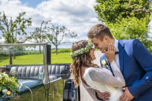 Braut und Bräutigam teilen einen freudigen Hochzeitsmoment neben einem Oldtimer. Die Braut trägt einen Blumenkranz und der Bräutigam einen blauen Anzug. Sie lächeln sich an und berühren ihre Stirn. Eine üppige grüne Landschaft und ein blauer Himmel mit Wolken dienen als Kulisse.