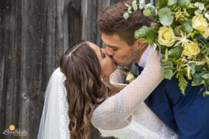 Eine Braut in einem weißen Spitzenkleid und Schleier küsst einen Bräutigam im blauen Anzug, der einen Strauß mit gelben Rosen und Grünpflanzen hält. Sie stehen vor einer hölzernen Kulisse und schaffen eine romantische Hochzeitsszene, die durch eine durchdachte Hochzeitsplanung perfekt eingefangen wurde.