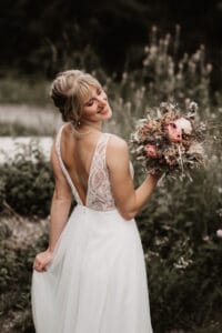 Eine Braut in einem ärmellosen Spitzenbrautkleid lächelt, während sie einen Strauß rosa und weißer Blumen hält und den perfekten Moment ihrer Hochzeit verkörpert. Sie steht im Freien, umgeben von Grün, und hat der Kamera leicht den Rücken zugewandt.