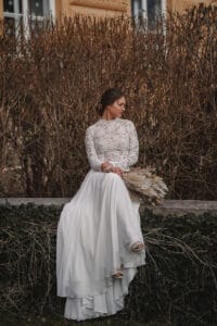 Eine Frau in einem langärmeligen Spitzenbrautkleid sitzt auf einem Steinsims und hält einen Strauß getrockneter Blumen in der Hand. Sie blickt zur Seite und betrachtet ihre Hochzeit inmitten kahler Sträucher und Weinreben. Im Hintergrund ist ein Gebäude mit großen Fenstern zu sehen, das diesem ruhigen Moment eine elegante Note verleiht.