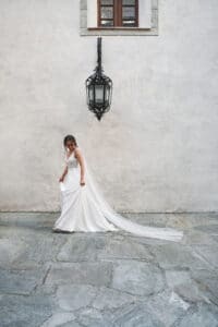 Eine Braut in einem fließenden weißen Kleid mit langem Schleier geht auf einem Steinpflaster und verkörpert die Eleganz der Hochzeitsplanung. Sie steht neben einer strukturierten weißen Wand mit einer großen, verzierten schwarzen Laterne, und darüber vervollständigt ein kleines Fenster diese bezaubernde Hochzeitsszene.