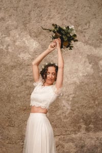 Eine Frau in einem Spitzentop und einem langen Rock hält einen Strauß Grünpflanzen über ihrem Kopf und verkörpert damit die Freude einer Hochzeit. Ihr lockiges Haar fällt wunderschön herab, während sie vor einem strukturierten, neutralen Hintergrund steht und die Essenz einer Hochzeit einfängt.