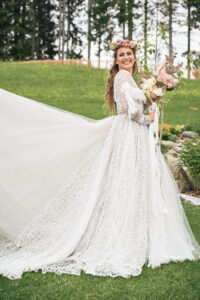 Eine Braut in einem Spitzenhochzeitskleid steht lächelnd mit Blumenkrone und Blumenstrauß im Gras. Ihr langer Schleier ist hinter ihr ausgebreitet und verkörpert die Essenz der Hochzeit inmitten von Bäumen und Grün im Hintergrund.