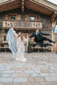 Eine Braut in einem weißen Spitzenkleid wirbelt ihren Schleier, während der Bräutigam im schwarzen Anzug verspielt neben ihr herumspringt. Sie stehen vor einer rustikalen Holzhütte mit Balkonen und schaffen eine fröhliche und skurrile Hochzeitsszene.