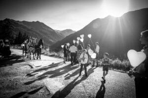 Ein Schwarzweißfoto zeigt eine fröhliche Gruppe, darunter Kinder, die herzförmige Luftballons halten. Sie haben sich draußen auf einer Straße versammelt, mit Bergen im Hintergrund – eine perfekte Szene zum Heiraten. Die Sonne wirft lange Schatten und links sorgt eine Pferdekutsche für einen Hauch zeitloser Romantik.