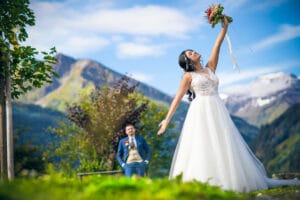 Eine Braut streckt freudig ihren Blumenstrauß vor einer Bergkulisse und einem klaren blauen Himmel in die Höhe und verkörpert damit den perfekten Moment ihrer Hochzeitsplanung. Sie trägt ein weißes Kleid, während ihr Bräutigam in einem blauen Anzug lächelnd inmitten üppigen Grüns steht.