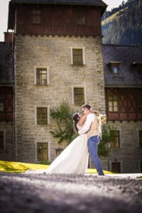 Ein Paar in Hochzeitskleidung umarmt sich vor einem großen Steingebäude mit grünen Fensterläden und verkörpert damit sein Hochzeitsglück. Die Braut hält einen Blumenstrauß, während das Sonnenlicht die Szene erhellt, während sich im Hintergrund anmutig Bäume und Hügel erstrecken.