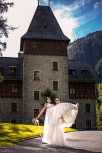 Ein Bräutigam trägt seine Braut freudig vor einem großen Steingebäude mit einem hohen Turm und fängt damit die Essenz ihrer Hochzeit ein. Die Braut hält einen Blumenstrauß und beide lächeln unter dem klaren blauen Himmel. Üppige grüne Bäume und Gras umgeben die Szene und schaffen eine malerische Kulisse.