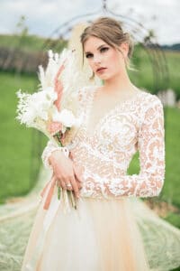 Eine Braut in einem eleganten, mit Spitze bestickten Brautkleid hält einen Strauß weißer und rötlicher Blumen. Sie steht in einem üppigen grünen Garten mit einem angedeuteten Hochzeitsbogen im Hintergrund. Ihr Haar ist zu einer lockeren Hochsteckfrisur gestylt.