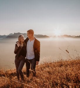 Ein Paar geht bei Sonnenuntergang Arm in Arm auf einem grasbedeckten Hügel spazieren, mit Bergen und Nebel im Hintergrund. Sie wirken glücklich, träumen von ihrer Hochzeit und genießen die friedliche Landschaft und das warme Licht.
