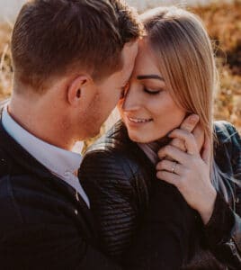 Ein Paar sitzt eng beieinander und teilt einen intimen Moment. Ihre Stirnen berühren sich, während sie sanft lächeln. Die Frau, die vielleicht über ihre Hochzeitsplanung nachdenkt, trägt einen Ring an ihrer linken Hand und eine schwarze Jacke. Der sanft verschwommene Hintergrund hüllt sie in warme Töne.