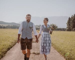 Ein Paar in traditioneller bayerischer Kleidung, das wahrscheinlich seine Hochzeit plant, schlendert Hand in Hand einen ländlichen Weg entlang. Der Mann trägt Lederhosen und eine Weste, während die Frau ein Dirndl trägt und einen Blumenstrauß hält. Sie sind von Feldern mit Bergen im Hintergrund umgeben.