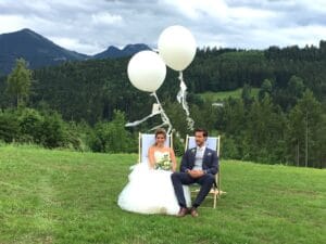 Braut und Bräutigam sitzen auf Stühlen in einer malerischen Umgebung im Freien, perfekt für eine Traumhochzeit, mit Bergen und üppigem Grün. Die Braut hält weiße Luftballons und beide sind formell gekleidet. Der bewölkte Himmel trägt zur ruhigen Landschaft bei, ideal für diejenigen, die ihre Hochzeitsreise planen.