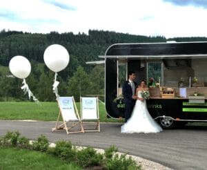 Ein frisch verheiratetes Paar steht neben einem eleganten schwarzen Food Truck im Freien und verkörpert damit ihren einzigartigen Hochzeitsstil. Zwei weiße Luftballons sind an Schildern in der Nähe befestigt und verleihen dem Ganzen eine festliche Note. Die Szene ist von üppigem Grün und sanften Hügeln unter einem bewölkten Himmel umgeben und fängt die Essenz ihrer Hochzeitsplanung ein.