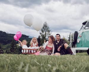 Eine Familie sitzt auf einer Wiese neben einem Oldtimer und genießt den Tag. Zwei Kinder halten Luftballons, einen weißen und einen rosa, die an eine fröhliche Hochzeit erinnern. Die Eltern kuscheln mit ihrem kleinen Hund, während Bäume und ein bewölkter Himmel die idyllische Kulisse bilden, perfekt für unvergessliche Hochzeitsmomente.