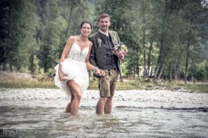 Ein Paar steht in einem seichten Fluss und hält sich an den Händen. Die Frau trägt ein kurzes weißes Kleid und der Mann ist in traditioneller Kleidung mit Jacke und knielangen Shorts gekleidet. Ihr heiterer Moment vor der Kulisse eines üppigen Waldes fängt die Essenz des Heiratens ein, glückselig umarmt von der Natur.