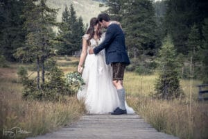 Eine Braut im weißen Kleid und ein Bräutigam in traditioneller Kleidung verbringen einen Moment auf einem Holzweg auf einer üppigen Wiese, umgeben von immergrünen Bäumen. Die Braut hält einen Blumenstrauß in der Hand, der die ruhige Atmosphäre ihrer Hochzeit verstärkt und die romantische Atmosphäre wirklich unvergesslich macht.