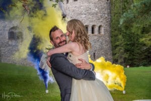 Ein Paar im Hochzeitskleid umarmt sich freudig vor einem Steinschloss. Im Hintergrund wabern gelbe und blaue Rauchwolken, die einen Kontrast zum üppigen Grün bilden und die Magie ihrer Hochzeit einfangen.