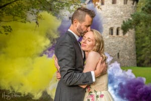 Ein Paar umarmt sich liebevoll vor einem burgähnlichen Steinturm, umgeben von gelben und violetten Rauchwolken. Während sie ihre Hochzeit planen, küsst der Mann im grauen Anzug sanft die Stirn der Frau. Sie trägt ein Kleid mit Blumenmuster und lächelt mit geschlossenen Augen.
