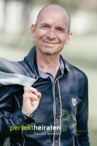 Ein Mann in einem blauen Hemd hält eine Jacke über der Schulter und lächelt in die Kamera. Der Text darunter lautet „perfektheiraten Harald Winkler“ und die grüne Silhouette eines Paares in Hochzeitskleidung fängt die Essenz exquisiter Hochzeitsplanung ein. Der Hintergrund ist leicht verschwommen.