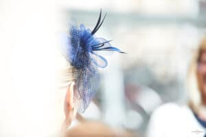Von hinten ist der Kopf einer Frau zu sehen, geschmückt mit einem eleganten blauen Fascinator mit Netz- und Federdetails, perfekt für eine Hochzeit. Der Hintergrund ist sanft verschwommen.