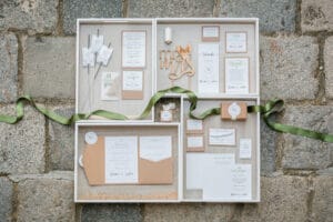 In einer Vitrine sind verschiedene Hochzeits-Briefpapiere ausgestellt, darunter Einladungen, Antwortkarten und Anhänger mit handgeschriebenem Text. Ein dekorativer hölzerner „Mr & Mrs“-Ausschnitt und eine grüne Schleife sind ebenfalls Teil des Arrangements, das auf einem Steinhintergrund platziert ist – perfekt für Ihre Hochzeitsplanung.