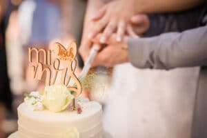 Hände eines Paares schneiden eine Hochzeitstorte an, die mit einem „Mr & Mrs“-Aufsatz und einer weißen Rose geschmückt ist. Die Torte ist elegant mit kleinen Blumen und grasähnlichen Akzenten dekoriert und spiegelt die Essenz sorgfältiger Hochzeitsplanung wider.