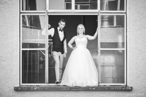 Ein Paar posiert in einem großen Fenster und feiert seine Hochzeit. Der Bräutigam trägt Weste und Fliege, während die Braut in ihrem trägerlosen Brautkleid umwerfend aussieht. Beide lächeln herzlich in die Kamera, eingerahmt von einer industriellen Kulisse mit glatten Metalloberflächen.