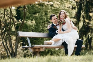 Ein fröhliches Paar im Hochzeitskleid sitzt zusammen auf einer Holzbank in einem üppigen Park. Die Braut, die einen Blumenstrauß hält, lacht, während der Bräutigam seinen Arm um sie legt. Sonnenlicht fällt durch die Bäume und schafft eine warme und romantische Atmosphäre, perfekt zum Heiraten.