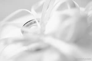 Ein Silberring mit eingraviertem Datum ist in weichen, weißen Stoff eingebettet, der mit zarten Bändern geschmückt ist. Das Bild strahlt Eleganz und Feierlichkeit aus und symbolisiert möglicherweise eine Hochzeit oder Hochzeitsplanung für einen besonderen Anlass.