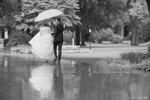 Braut und Bräutigam gehen an einem regnerischen Tag in einem Park spazieren, wobei der Bräutigam einen Regenschirm über sie hält. Ihr Spiegelbild schimmert auf dem nassen Weg und macht den Moment ihrer Hochzeit perfekt. Bäume und ein Laternenpfahl stehen sanft im Hintergrund des zeitlosen Schwarzweißfotos.
