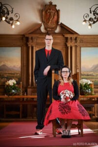 Ein Paar posiert elegant in einem formellen Rahmen und fängt die Essenz ihrer Hochzeit ein. Der Mann steht in einem schwarzen Anzug mit rotem Hemd, während die Frau anmutig auf einem Holzstuhl sitzt, in ihr rotes Kleid gekleidet und einen Blumenstrauß in der Hand. Aufwändige Holzarbeiten und ein exquisites Gemälde verleihen diesem intimen Moment Charme.