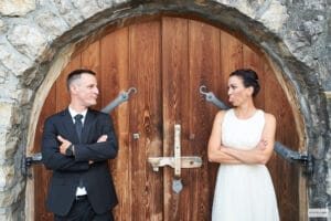 Ein Mann im schwarzen Anzug und eine Frau im weißen Kleid stehen sichtlich in der Freude ihrer Hochzeitsplanung versunken vor einer großen Holztür. Die Frau streckt dem Mann, der mit verschränkten Armen amüsiert wirkt, spielerisch die Zunge heraus. Beide lächeln leicht.