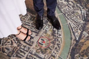 Ein Paar Füße steht auf einer detaillierten Luftbildkarte und verkörpert den Ablauf der Hochzeitsplanung. Die linke Person trägt elegante schwarze Riemchensandalen, aus denen ein weißes Kleid hervorschaut, während die rechte Person glänzende schwarze Abendschuhe trägt, die zu dunklen Hosen passen. Ein roter Kreis hebt einen Orientierungspunkt auf der Karte hervor und symbolisiert zukünftige Feierlichkeiten.