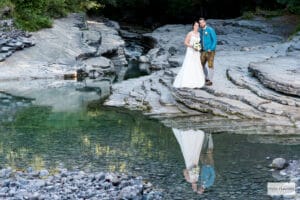 Braut und Bräutigam stehen auf felsigem Gelände neben einem reflektierenden Wasserbecken in einem Waldgebiet. Die Braut trägt ein weißes Kleid und hält einen Blumenstrauß, während der Bräutigam, der den Geist der Hochzeit verkörpert, eine türkisfarbene Jacke und braune Hosen trägt. Ihr Spiegelbild schimmert wunderschön im Wasser.