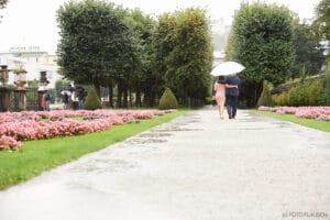 Ein Paar geht Arm in Arm unter einem Sonnenschirm durch einen Garten und stellt sich seine Hochzeit vor. Rosa Blumen säumen den Weg, und zu beiden Seiten stehen große Bäume. Im Hintergrund sind weitere Menschen zu sehen, ebenfalls mit Sonnenschirmen, an diesem romantischen Regentag, der sich perfekt für die Hochzeitsplanung eignet.