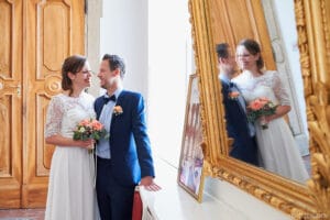 Ein Paar im Hochzeitskleid lächelt sich an, während es vor einem großen, verzierten Spiegel steht und die Freude des Heiratens verkörpert. Die Braut hält einen Blumenstrauß und die Holztüren und der goldene Rahmen verleihen dieser bezaubernden Hochzeitskulisse Eleganz.