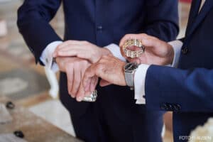 Zwei Personen in Anzügen gestikulieren mit ihren Händen und halten in einem formellen Rahmen jeweils einen verzierten Ring. Die Armbanduhr einer Person ist sichtbar und verleiht diesem Hochzeitsmoment einen Hauch von Eleganz.