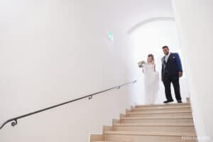 Eine Braut im weißen Kleid und ein Mann im dunklen Anzug steigen anmutig gemeinsam eine hell erleuchtete Treppe hinab, die Frau hält einen Blumenstrauß in der Hand. Über dem Torbogen oben hängt ein Notausgangsschild, das diesen bezaubernden Hochzeitsmoment subtil umrahmt.