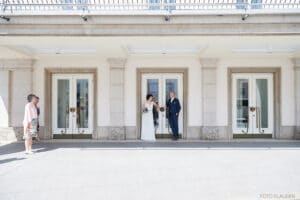Braut und Bräutigam stehen vor einem weißen Gebäude mit großen Glastüren und verkörpern die Freude am Heiraten. Die Braut hält einen Blumenstrauß, während ein Paar in der Nähe diesen hell erleuchteten, feierlichen Moment im Freien beobachtet. Die Szene spiegelt durchdachte Hochzeitsplanung in ihrer schönsten Form wider.