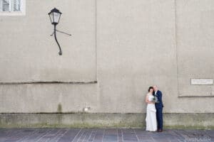 Braut und Bräutigam stehen dicht beieinander vor einer großen, strukturierten beigefarbenen Wand, über der eine schiefe Straßenlaterne hängt. Sie feiern ihre Hochzeit, die Braut hält einen Blumenstrauß und beide lächeln. Der Boden besteht aus rechteckigen Fliesen und eine kleine Plakette verleiht diesem perfekten Rahmen zum Heiraten Charme.