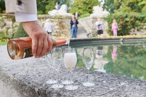 Eine Person gießt Champagner in ein Glas neben drei leeren Gläsern auf einer Steinfläche und bereitet damit die Bühne für eine elegante Hochzeit. Im Hintergrund versammeln sich die Gäste an einem ruhigen Teich, umgeben von üppigem Grün und Skulpturen, was die Kunst der Hochzeitsplanung perfekt verkörpert.