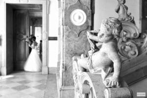 Im Vordergrund steht eine steinerne Cherub-Statue, die auf eine große Treppe blickt. Im Hintergrund steht eine Braut in einem fließenden Kleid neben einer offenen Tür, die vom einfallenden Licht sanft beleuchtet wird. Die Szene verkörpert die elegante Gelassenheit der Hochzeitsplanung und fängt zeitlose Momente der Freude und Liebe ein.
