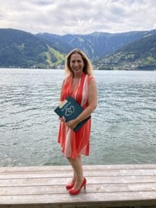 Eine Frau in einem rot-weiß gestreiften Kleid steht auf einem Holzsteg am See und hält einen Ordner mit einem Herzmotiv in der Hand, vielleicht etwas zum Thema Hochzeitsplanung. Sie lächelt herzlich, eingerahmt von Bergen und einer malerischen Stadt im Hintergrund. Es ist ein bewölkter Tag.