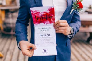 Eine Person im blauen Anzug hält eine Hochzeitseinladung mit roten Aquarellakzenten in der Hand, auf der das Hochzeitsdatum 18.02.17 steht. Eine rote Blume ist an ihrem Revers befestigt, während sie auf einem Holzdeck steht und die Essenz einer durchdachten Hochzeitsplanung verkörpert.