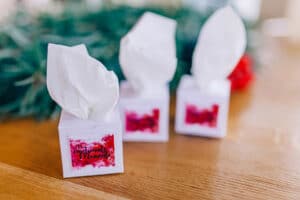 Drei kleine Taschentuchboxen mit herausstehenden weißen Taschentüchern stehen auf einer Holzoberfläche, perfekt für die emotionalen Momente während der Hochzeit. Die Boxen sind mit Etiketten mit einem abstrakten Design und Text in Rot und Rosa versehen, vor einem verschwommenen grünen und roten Hintergrund, der die festliche Atmosphäre ergänzt.