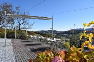Außenterrasse mit weißen Stühlen und Tischen auf einem Holzdeck, perfekt für eine Hochzeitsfeier, mit Blick auf eine malerische Landschaft. Die Aussicht umfasst sanfte Hügel unter einem klaren blauen Himmel. Im Vordergrund sind bunte Blätter und Kies zu sehen, ideal für zauberhafte Hochzeitsplanungserinnerungen.