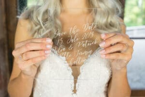Eine Person mit langem, welligem Haar hält eine durchsichtige Acrylkarte mit eleganter Handschrift: „Save the Date 15. Juli 2018 Julia & Patrick“, ein Vorgeschmack auf ihre bevorstehende Hochzeit. Die manikürten Fingernägel der Person ergänzen die Eleganz ihres weißen Spitzengewandes.