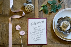 Ein Hochzeitsmenü mit eleganter Kalligraphie liegt auf einem Holztisch und verkörpert einen perfekten Hauch von Hochzeitsplanung. Es gibt ein rosa Band, natürliche Dekorationen wie Samenkapseln, grüne Blätter und einen Teller mit einer Kerze. Daneben liegen zwei Stäbchen mit „Ja“ auf rosa Kreisen.